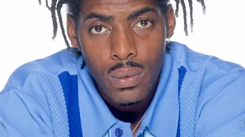 Rapper Coolio poses in a blue shirt against a white background.