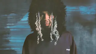 Yung Kayo poses in front of a blue shaded backdrop in a fur hat, looking directly at the camera