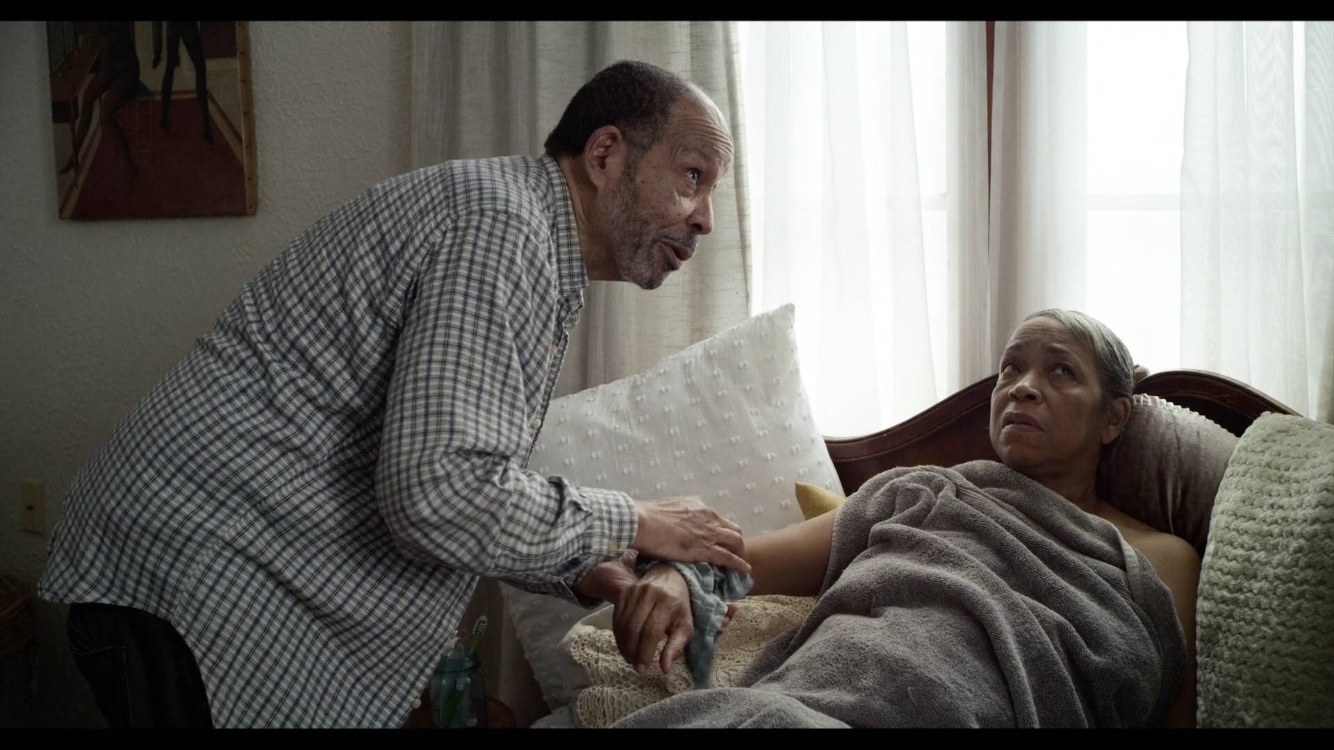 A man in a flannel shirt checks holds the wrist of a woman sitting down underneath a blanket.