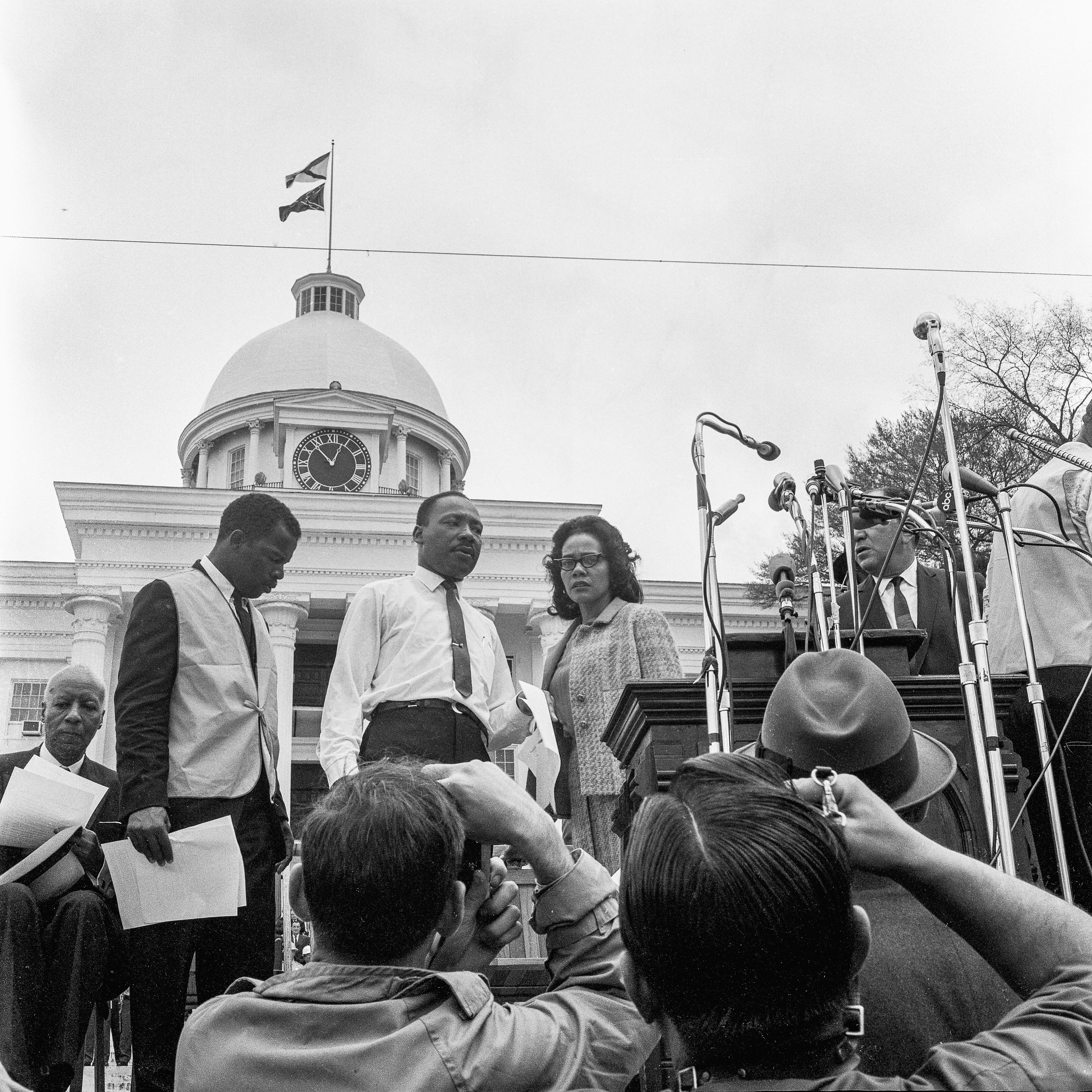 View of American religious and Civil Rights leaders John Lewis (in vest) and Martin Luther King Jr (1929 - 1968) and his wife, Coretta Scott King (1927 - 2006), on the podium before the Selma to Montogomery March rally on the steps on the Alabama State Capitol, Montgomery, Alabama, March 25, 1965. Also visible is union leader A Philip Randolph (1889 - 1979) (seated at left). The Confederate and Alabama flags fly over the Capitol. (Photo by Charles Shaw/Getty Images)