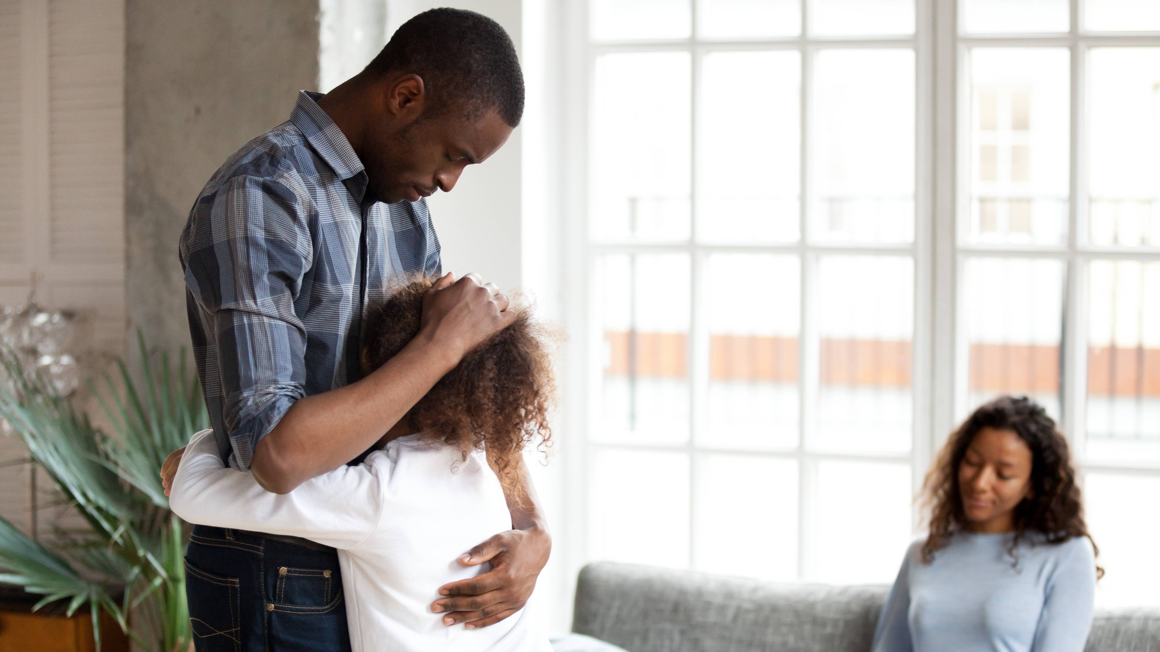 African American family in living room at home, sad father embracing little preschooler daughter as mom sits on the couch looking upset. 