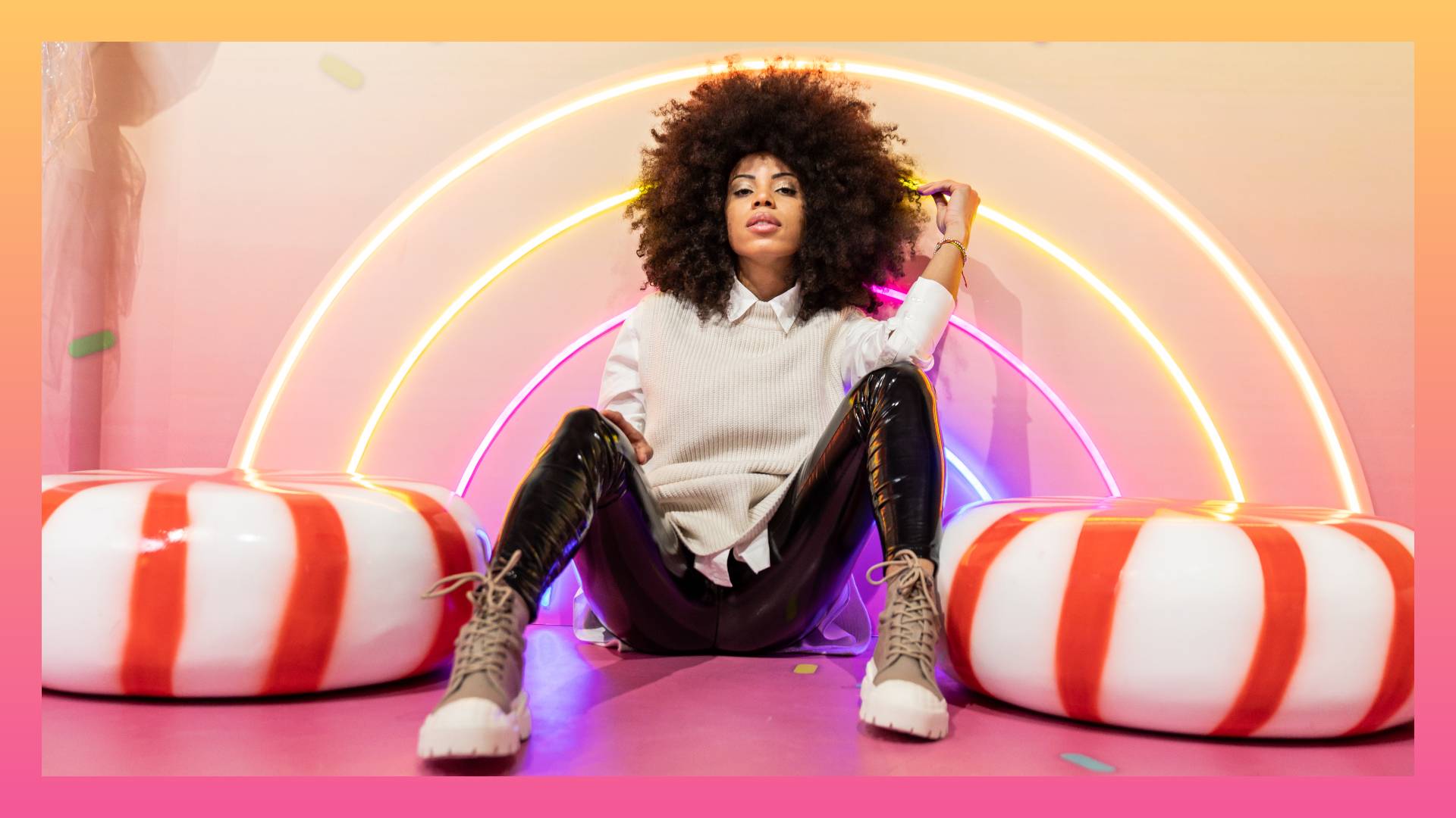 Portrait of young afro woman sitting next to a lollipop and huge candies in a studio. Sugar and sweets concept.