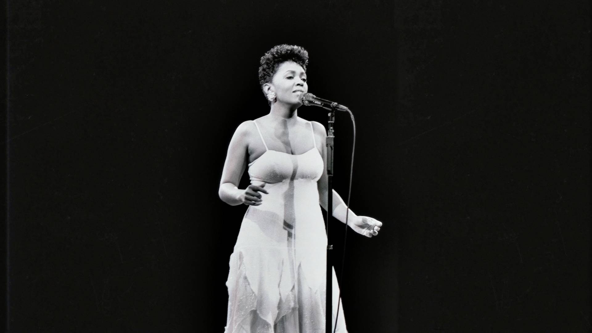 Legendary artist Anita Baker in an older black and white photo, performing on stage in a white dress.