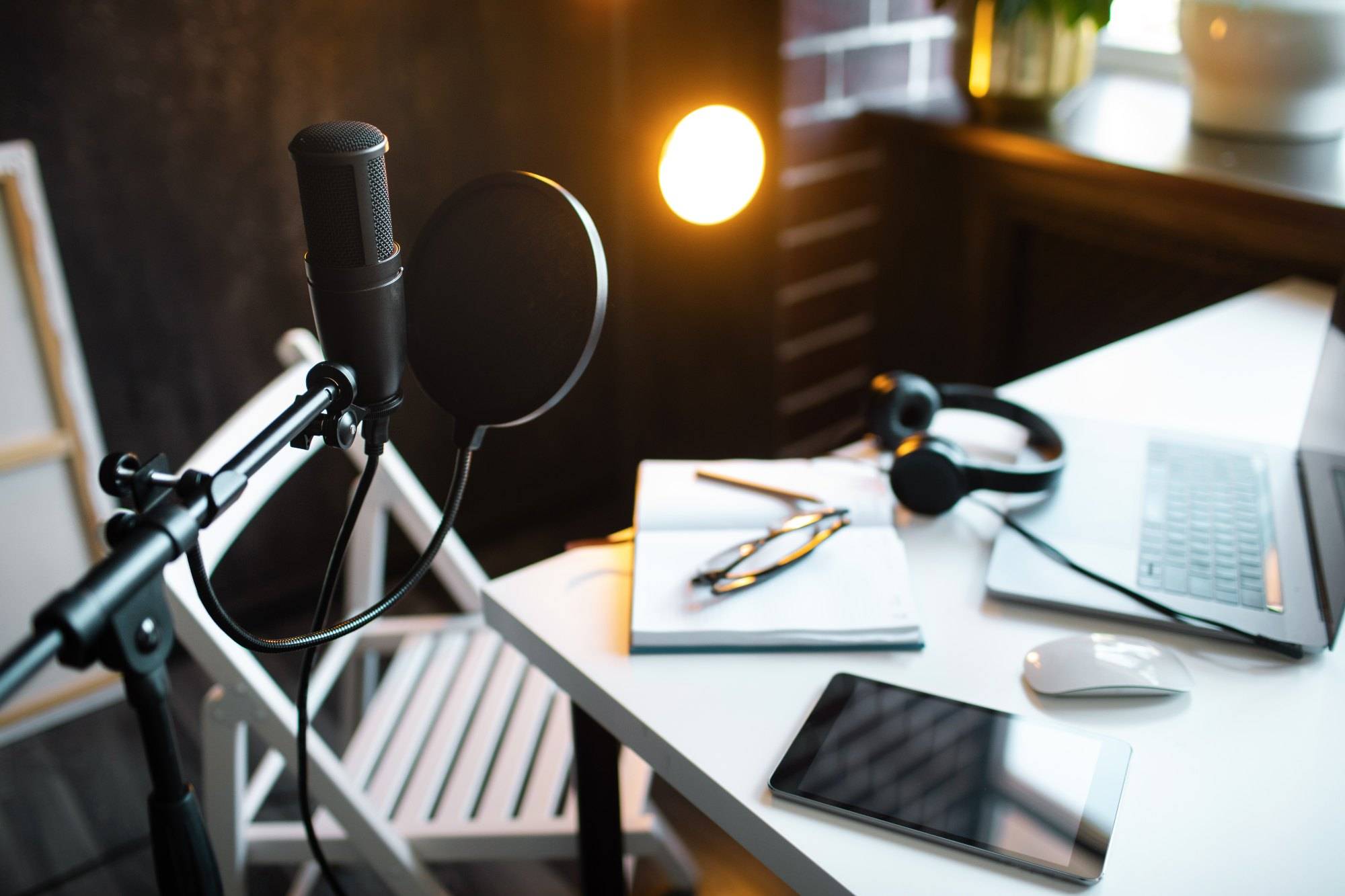 Podcast streaming at home. Audio studio with laptop, microphone with pop filter and headphones on white table against black wall with warm lights. Blogger concept.