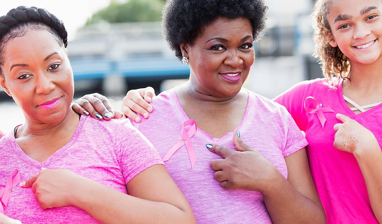 A group of three multi-ethnic women of mixed ages standing together at a breast cancer awareness event, wearing pink, pointing to the ribbons pinned to their shirts. The youngest, on the right, is a mixed race African-American and Caucasian teenage girl. The mature African-American woman in the middle is in her 50s and her friend is in her 30s.