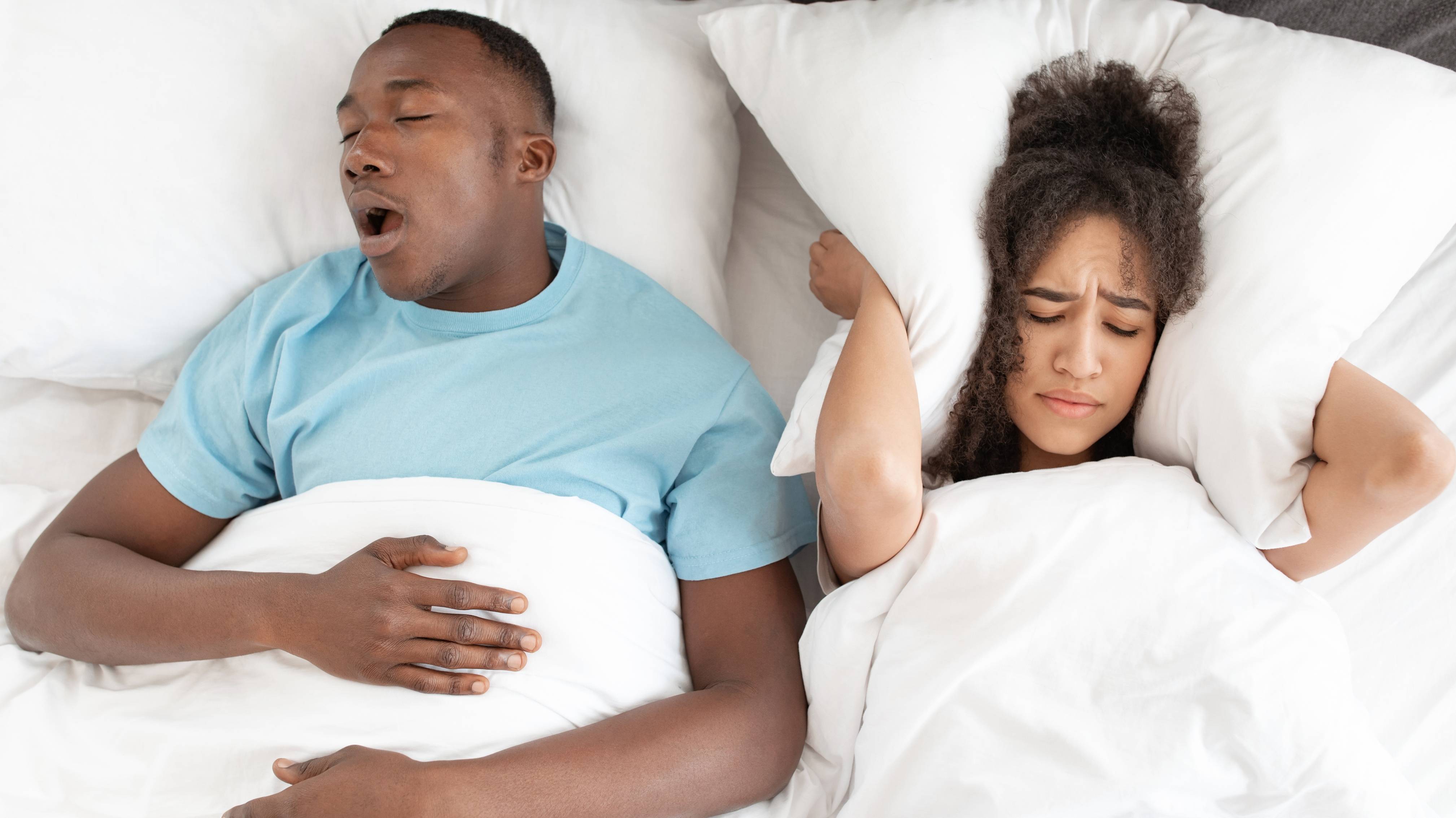 Young tired guy sleeping and snoring, next to sleepless woman covering her ears with pillow so as not to hear noise.